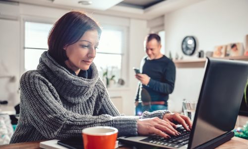 Woman wearing grey sweater using laptop at home office while her husband using smart phone in the background